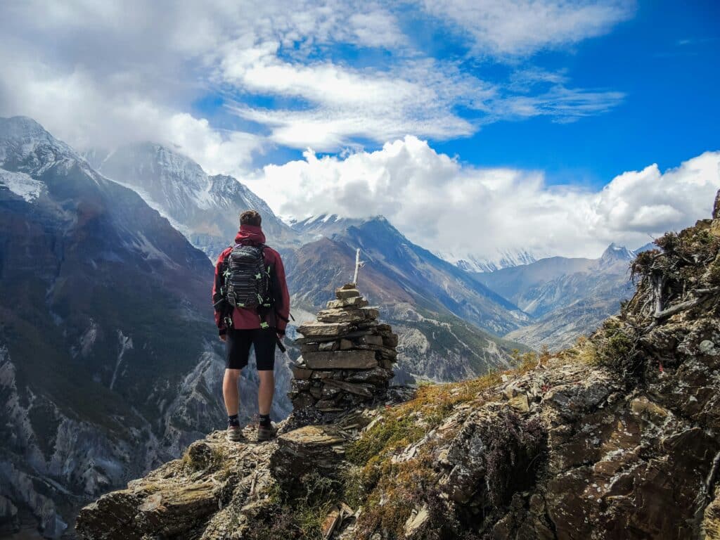 Um caminhante está em uma borda rochosa da montanha, viajando com propósito, com vista para os picos nevados e um vale abaixo.
